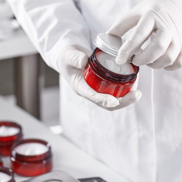 Factory worker in white gown and gloves holding red container with skin care product touching it with finger testing quality. Representing new product on factory. Showing high quality beauty mask
